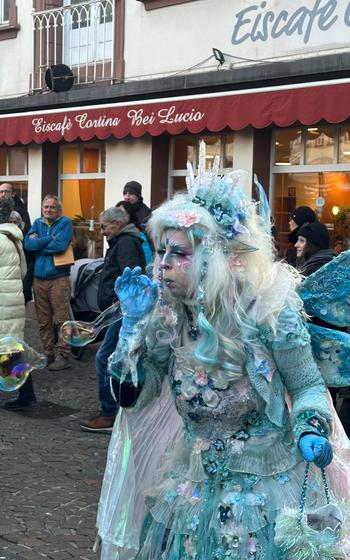 A close up of a blue fairy with an icy blue dress adorned with blue flowers, bright blue wings and a blue crystal tiara, blowing a big bubble from her wand. 