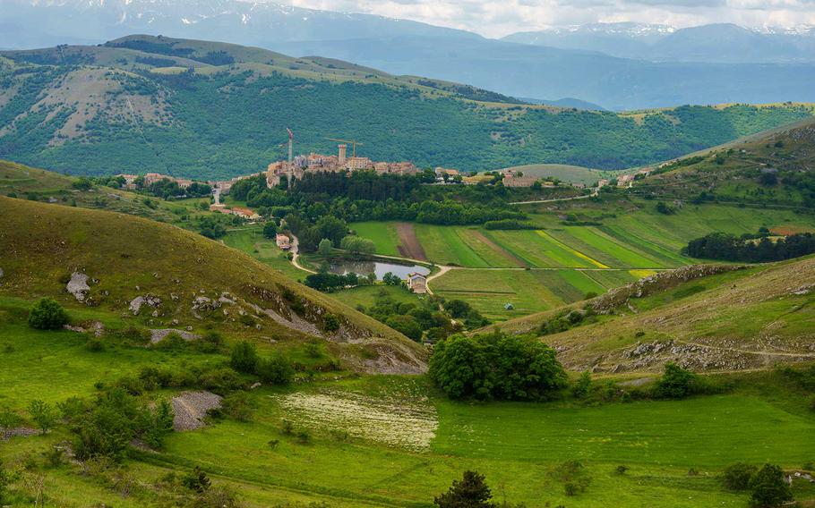 A breathtaking landscape of the Gran Sasso region in Italy, featuring rolling green hills, patchwork fields, and a charming medieval village set against the backdrop of the rugged mountains.