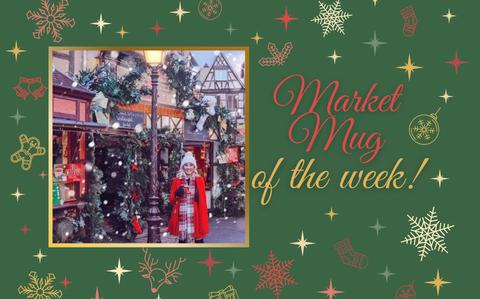 Photo Of A post-card style photo of a woman in a red coat holding a red, boot-shaped mug with the background of holiday-decorated half timber houses. 