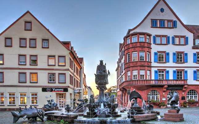 Two historical buildings stand behind a sculptural fountain with fish. One building has blue bright blue shutters