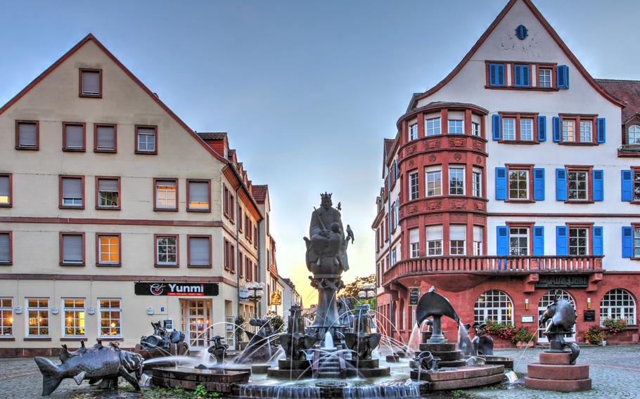 Two historical buildings stand behind a sculptural fountain with fish. One building has blue bright blue shutters