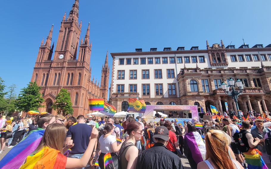 Crowd, facing away from camera, looking towards a stage, holding Pride flags and wearing Pride attire at the Wiesbaden Rathaus.