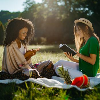 The blonde girl is reading the book while her friend is chatting and browsing via the mobile phone