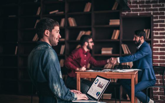sales Manager working on laptop with a marketing report on the background of business partners handshaking