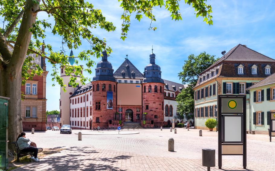 A picturesque square surrounded by historic buildings in Germany, featuring a red sandstone structure with two rounded towers and a central gable. The scene is framed by leafy trees under a clear blue sky, with a relaxed atmosphere created by the open plaza and scattered pedestrians. This charming location invites visitors to explore its architectural beauty and enjoy its tranquil setting.