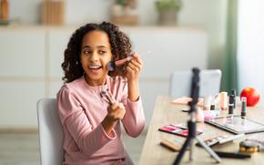 Beauty blogging. Cheerful african american teen recording cosmetics product review, showing various brushes and applying powder. Girl filming video for her channel using phone on tripod