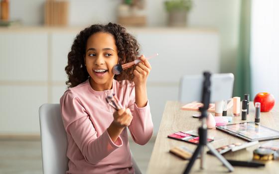 Beauty blogging. Cheerful african american teen recording cosmetics product review, showing various brushes and applying powder. Girl filming video for her channel using phone on tripod