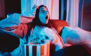 Close up portrait of young girl at home in a dark room with a popcorn bucket playing with her dog and sits in front of a monitor or TV.