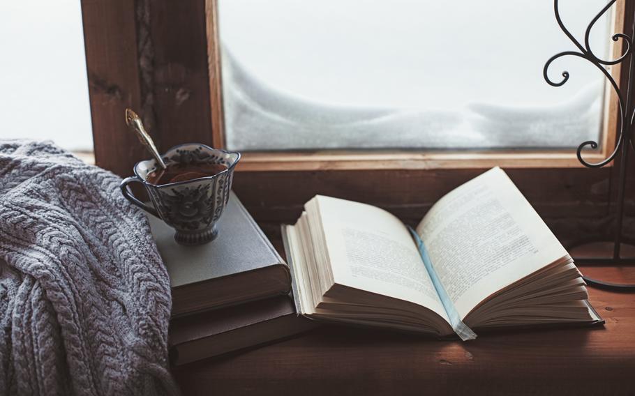 (From left to right) Folded sweater, cup of lemon tea and open book with sky blue ribbon book mark between the two pages on wooden window sill in old house. Behind the items is a window which reveals a snowy outside. 