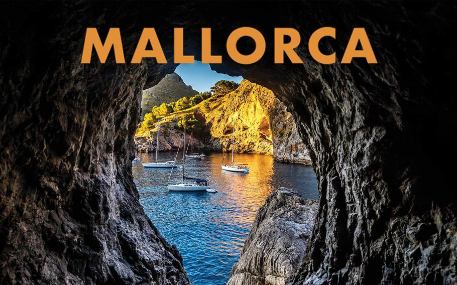 View of a picturesque bay in Mallorca, framed by the rocky entrance of a cave, featuring turquoise waters, anchored sailboats, and sunlit golden cliffs surrounded by lush greenery.