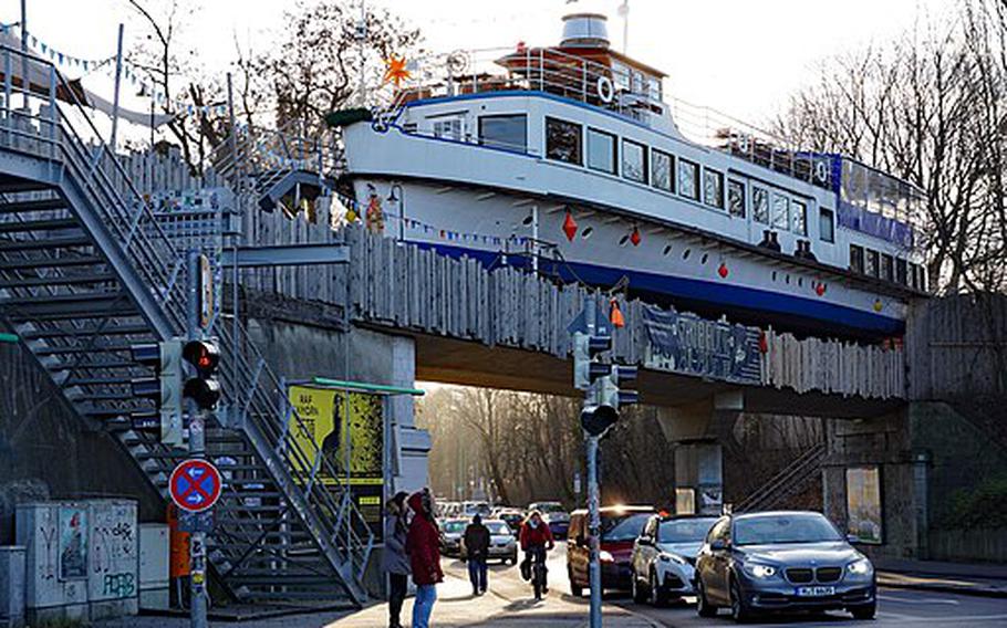 An up-close photo of a blue and white sailboat on a wooden bridge, cars drive by underneath. 