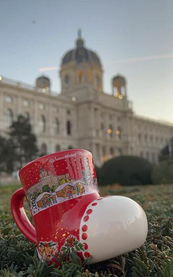 An up-close portrait of a red boot-shaped mug sittin gin the green grass. Behind it is domed building. 