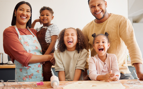 Family laughing as they bake in the kitchen | Table covered in flour and dough | From left to right behind the table: Mom holding a toddler on her hip, laughing young girl with eyes closed, dad smiling at camera, another young girl smiling with her mouth wide open