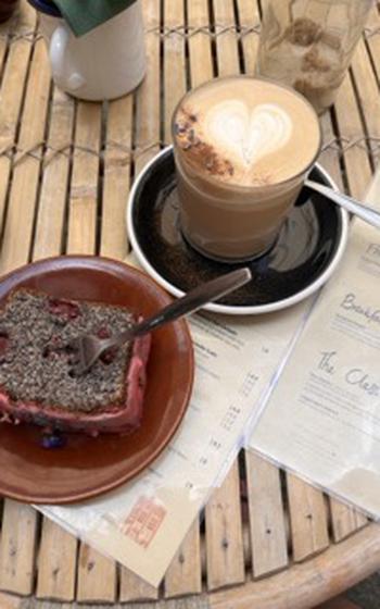 a slice of chocolatey-seedy cake with red frosting and a latte with a foam heart. 