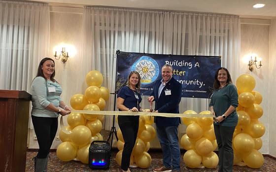 Photo Of Two women holding a large yellow ribbon with each woman on one end of the ribbon. A man and a woman are standing behind the ribbon at the center and the man is holding a large pair of scissors about to cut the ribbon.