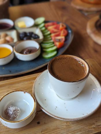 Turkish coffee in a cup with a piece of Turkish candy on a dish on the left of the cup. There is a blurred plate of fruit in the background.