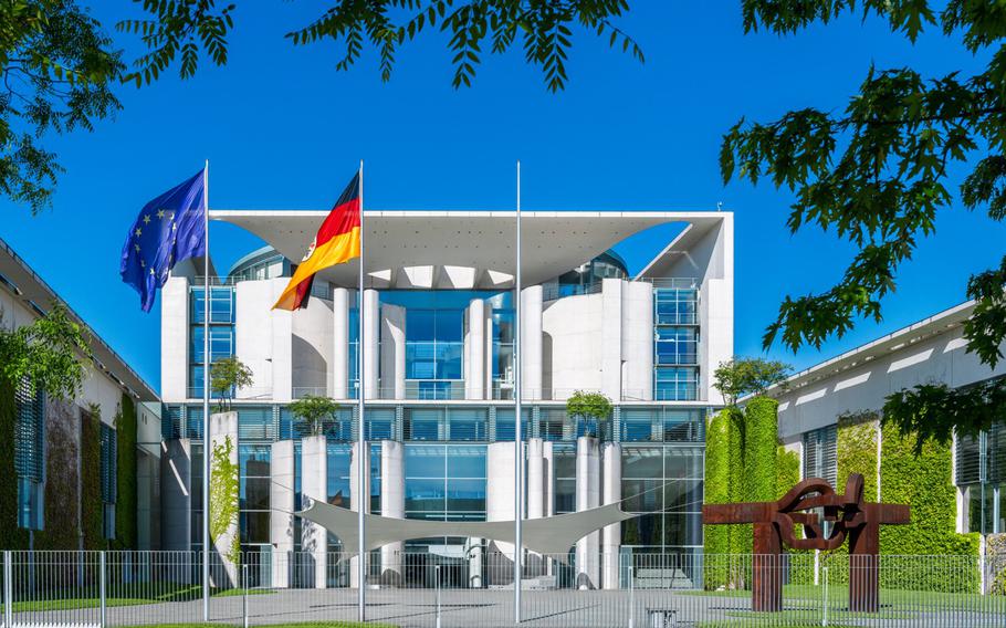 A tan and white building, the chancellery, with a german and EU flag flying high against a bright blue sky. 