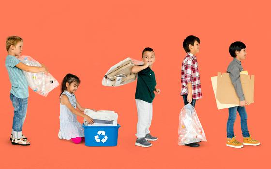 From left to right: Boy facing right holding clear bag of plastics, girl kneeling facing down at blue recycling bin, boy holding paper bags behind shoulder looking just behind the camera, boy facing right holding a clear bag of plastics and boy facing right holding flattened cardboard boxes.