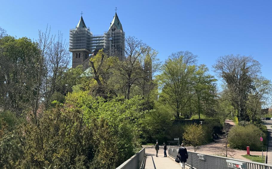 Church spires jut above bright green trees into a clear blue sky as pedestrians stroll towards it on a bridge. 