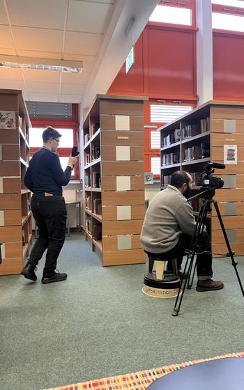 a student quietly films a video interview from behind library shelves