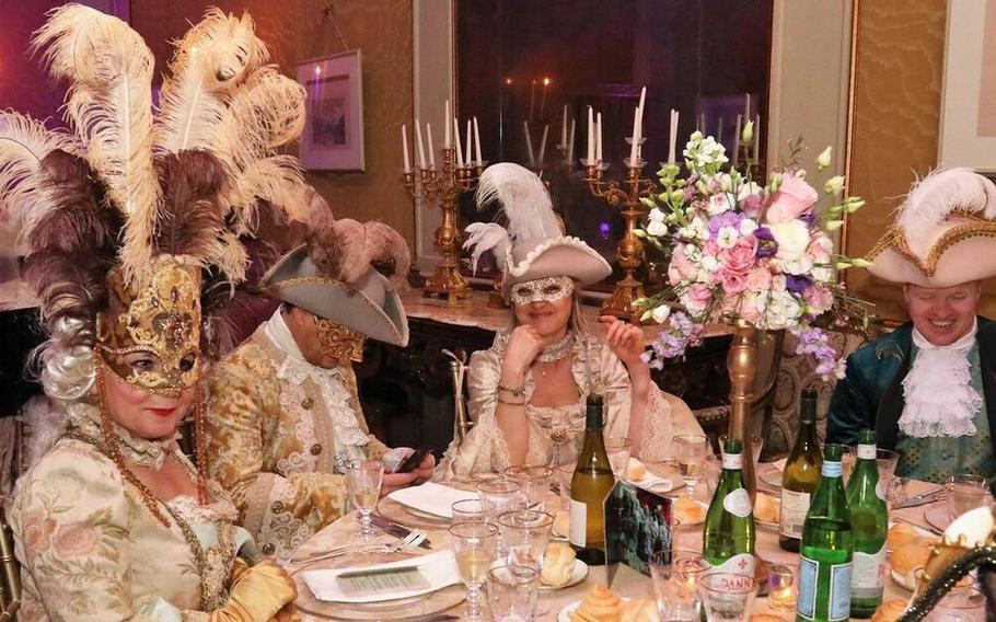 A lavish Grand Ball Carnival setting in Venice, Italy, featuring elegantly dressed guests adorned in opulent 18th-century-style attire and intricate Venetian masks. Feathered headdresses and ornate costumes reflect the grandeur and artistry of the Venetian tradition. The table is set with fine dining elements, including candelabras, floral arrangements, and bottles of wine, adding to the atmosphere of luxury and celebration in this timeless event.