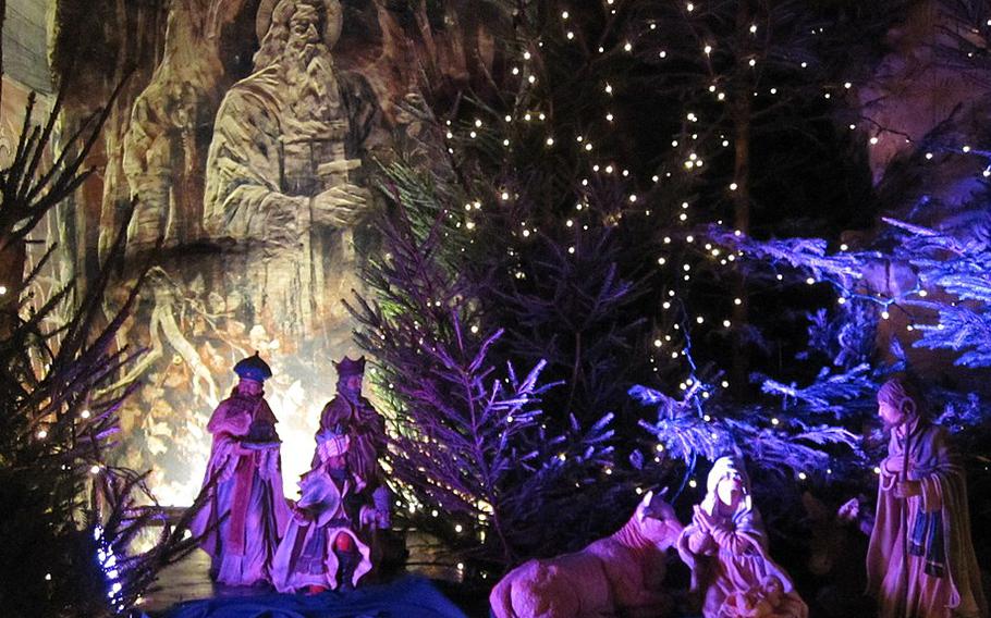 A purple and blue close up of a Nativity scene displayed in front of the cave wall withe evergreen trees in the background.