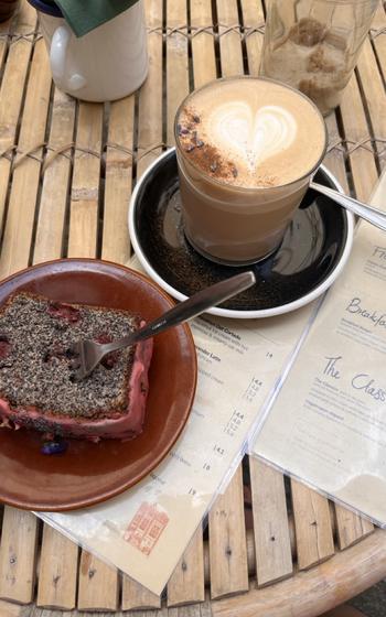 A slice of chocolatey cake with cherries in it and bright red frosting next to a latte with a heart. 