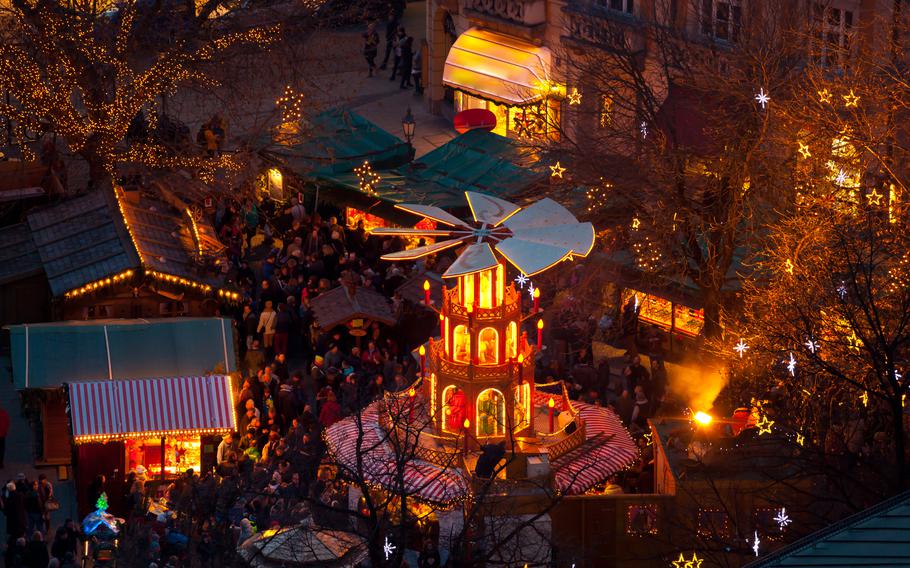 This photo captures a magical scene of a bustling Christmas market at twilight. The centerpiece is a traditional wooden Christmas pyramid carousel, beautifully illuminated with warm golden lights, creating a festive glow. Surrounding it are cozy market stalls with striped awnings and twinkling fairy lights, offering seasonal treats, handcrafted gifts, and holiday cheer. The trees are adorned with glowing star-shaped decorations, adding to the enchanting atmosphere. A cheerful crowd fills the market, enjoying the joyous ambiance of this holiday celebration. The backdrop of historic buildings completes the picturesque charm of this wintry wonderland.