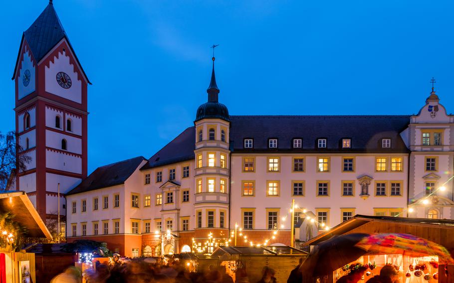 A large, white monastery building and tower are illuminated from below where a Christmas market is set up.