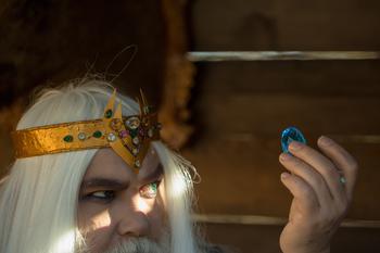 Old bearded man wizard in golden crown with long white hair and beard holding blue gem stone on wooden background