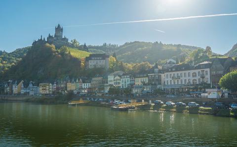 Photo Of view of the city of Cochem and castle, Germany from the Moselle River, with sun flare in the blue sky and reflecting on the river at the front on the photo