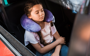 A young child sleeping in a car seat wearing a purple neck pillow