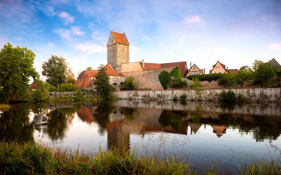 a far away photo of the medieval town of Dinkelsbuhl reflecting in the water. the tan building is surrounded by trees and a clear sky