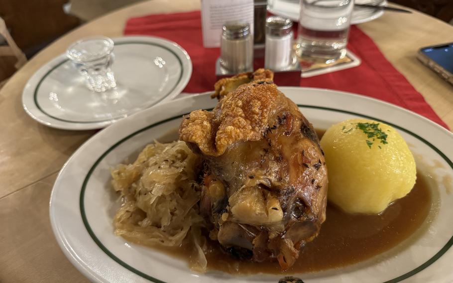 A plate with a crispy pork shoulder gravy, coleslaw, and potatoe dumpling, along with a clear schnapps. 