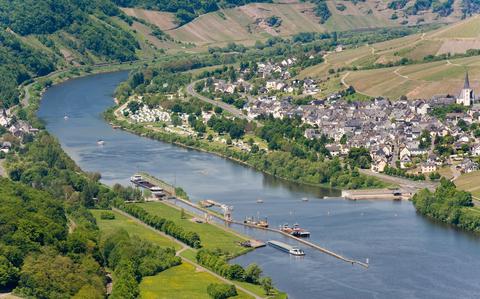 Photo Of Aerial view of Moselle valley. To the left are green trees and bushes. To the center is the Moselle river featuring several large boats in the water. To the right are buildings and churches. At the back are the wine vineyards.