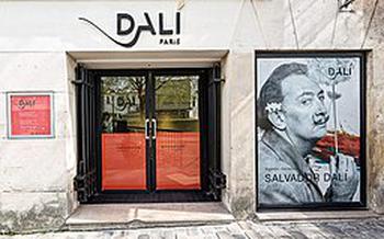 A pretty white building with and elegant font spelling “Dali Paris” there is a black and white picture of Salvador Dali with his iconic upturned mustache on the right by the doors. There are accents of red on the doors and in the picture. 