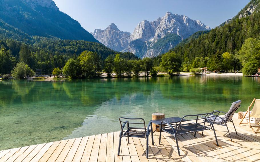 two chairs over look a clear mountain lake surrounded by lush green trees and mountains. 