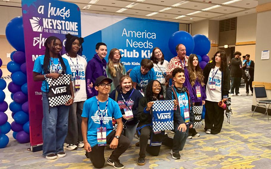 A group of teenagers poses in from of a blue display board in a conference center setting.