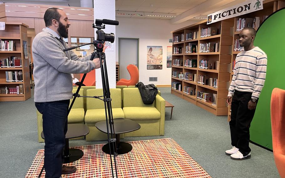 In a library a man is at a camera filming a student in front of a greenscreen