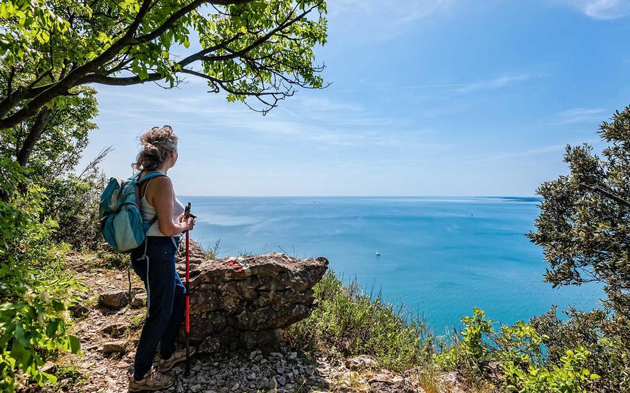 A hiker pauses along a scenic cliffside trail in the Tuscan Archipelago, taking in the stunning view of the turquoise Mediterranean Sea under a clear blue sky.