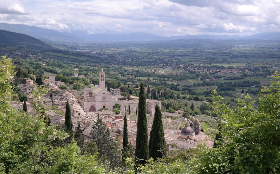 Far off view of Assisi Castle with two skinny and tall trees in front with the sky and mountains behind it.