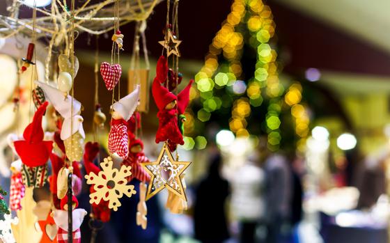 Several handmade Christmas ornaments hanging on a display. They include wooden snowflakes, angels, wooden stars. There is a blurred Christmas tree and crowd in the background.