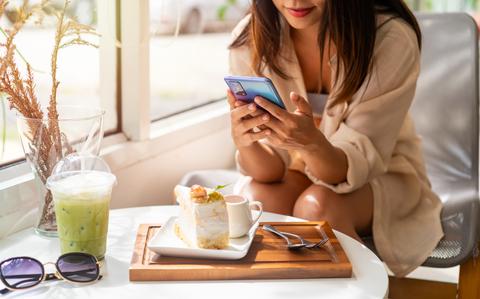 Photo Of Woman looking at smartphone.  She is sitting at a table with a piece of cake, iced green drink and sunglasses on the table.
