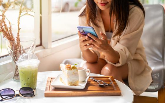 Woman looking at smartphone.  She is sitting at a table with a piece of cake, iced green drink and sunglasses on the table.