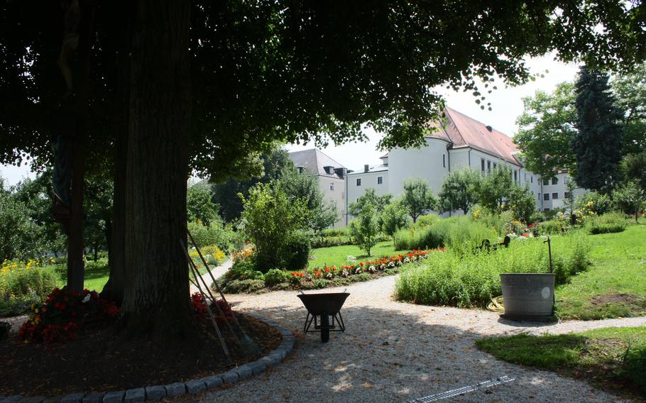 A wheelbarrow sits in the shade under a tree in a formal garden with a large white monastery building in the background. 