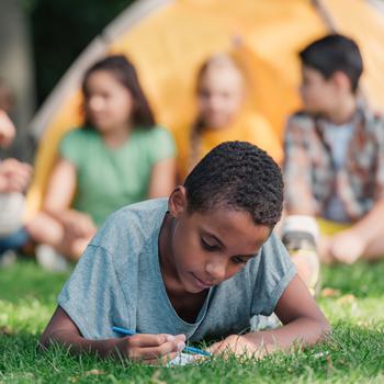 selective focus of happy african american boy writing in notebook near friends in camp