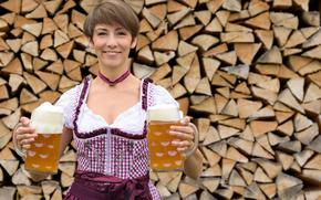Happy  woman in a dirndl holding two pints of beer in her hands as she stands in front of a rustic woodpile