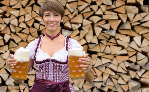Photo Of Happy  woman in a dirndl holding two pints of beer in her hands as she stands in front of a rustic woodpile
