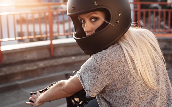 Young blonde haired  woman in leather jacket, wearing a black helmet is sitting on vintage custom motorbike