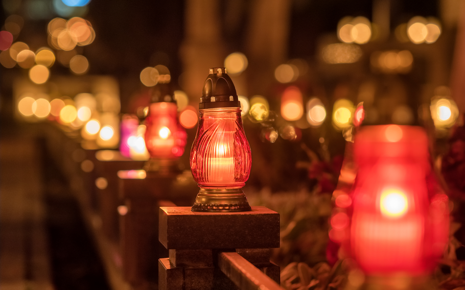 A row of glowing red and gold votive candles lines a stone railing, softly illuminating the surroundings with warm, gentle light. The blurred background features more candles, adding depth and a peaceful ambiance, symbolizing remembrance and reflection.